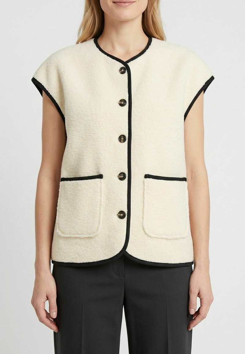 Femme portant un gilet sans manches en polaire crème avec bordure noire, cinq boutons devant et deux poches avant, assorti à un pantalon noir.