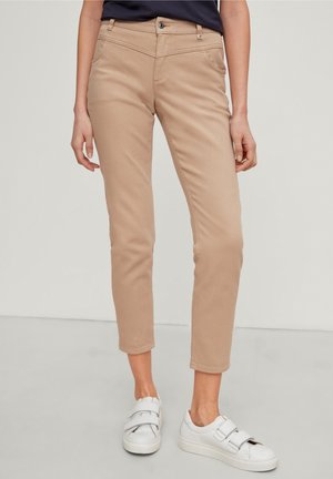 Osoba nosząca beżowe, dopasowane, skrócone spodnie oraz białe sneakersy na rzepy, stojąca na jasno szarej podłodze na jednolitym tle.