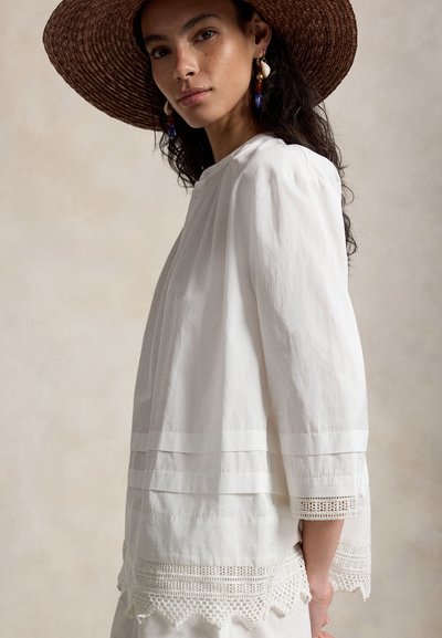 Γυναίκα που φοράει καπέλο με φαρδύ γείσο καφέ χρώματος, λευκό μπλουζάκι με μακριά μανίκια και δαντελένια διακόσμηση, καθώς και πολύχρωμα σκουλαρίκια με κοχύλια, κοιτάζοντας ελαφρώς προς τα αριστερά.