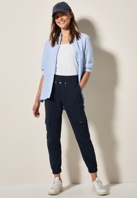 Blå skjorta med knappar över en vit linne, marinblå elastiska cargo-byxor med fickor och vita sneakers. Avslappnad stil med en keps.