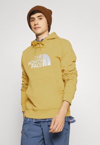 Moletim amarelo com um bolso tipo canguru e logótipo branco bordado. Combinado com uma camisa xadrez azul e um gorro de malha castanho. Textura suave.