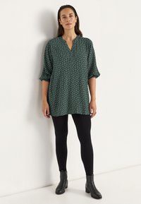 Groene tuniek met een V-hals en 3/4 mouwen, gecombineerd met zwarte legging en enkellaarsjes. De stof heeft een geometrisch patroon.