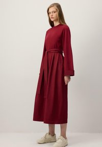 Bordové šaty s dlhými rukávmi, priliehavým živôtikom, opaskom na viazanie v páse a splývavou sukňou. Nosené so svetlými teniskami.