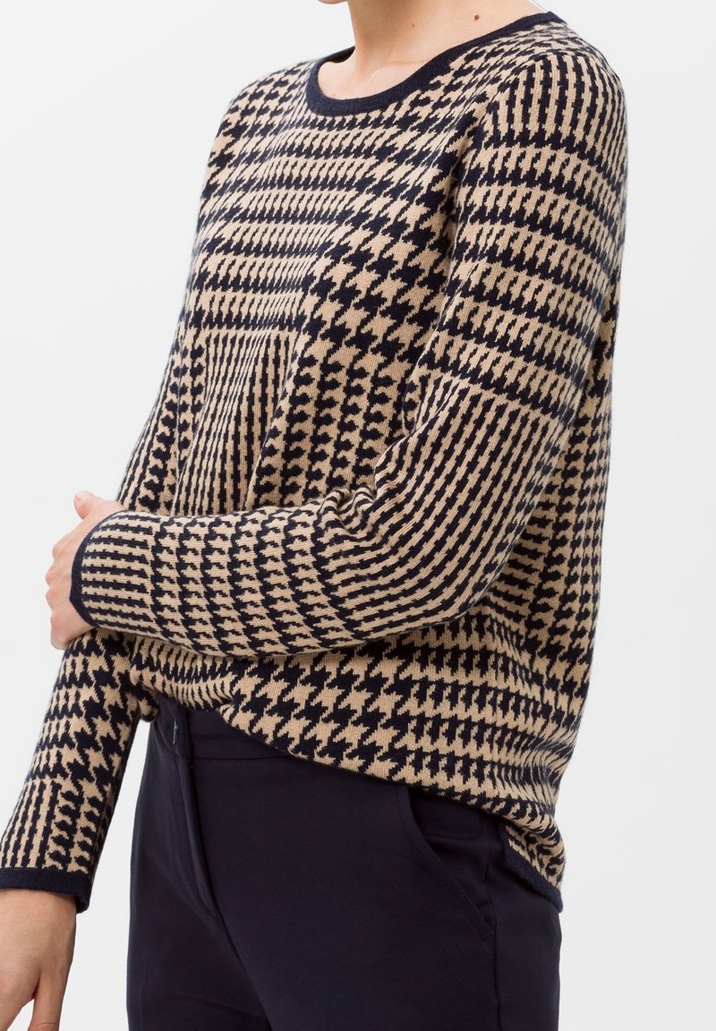 Femme portant un pull à manches longues beige et noir à motif pied-de-poule avec bordure noire et pantalon taille haute foncé, fond blanc.