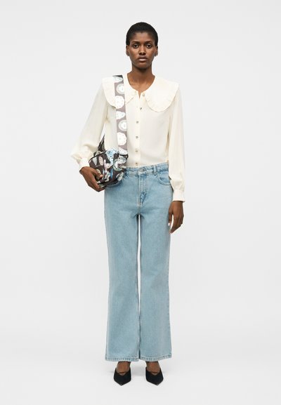 Γυναίκα που φοράει κρεμ μπλούζα με μεγάλο γιακά, ανοιχτό μπλε τζιν παντελόνι με φαρδιά μπατζάκια, μαύρα παπούτσια με μυτερή μύτη και τσάντα ώμου με σχέδια.