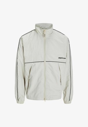 Chaqueta de crema ligera con cuello alto, cremallera frontal y franjas laterales negras; cuenta con puños elásticos y un logo discreto en la parte delantera.