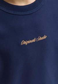 Navy sweatshirt with ribbed crew neck, featuring gold cursive embroidery that reads "Originals Studio" on the front. Soft texture, casual design.