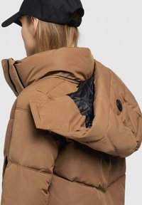 Braune gepolsterte Jacke mit hohem Kragen und verdecktem schwarzen Reißverschluss. Schwarze Innenauskleidung und ein Knopfdetail an der Kapuze.