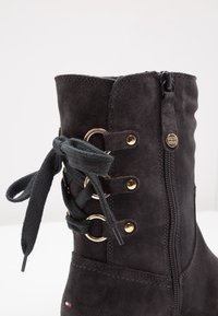 Stivale alla caviglia in suede nero con zip laterale, occhielli dorati e dettagli in pizzo a fiocco. Texture liscia con cuciture rinforzate.