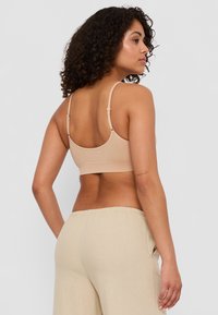 Beige trådlös BH med tunna band, mjuk yta och bårdformad design. Modellen bär ljusa, löst sittande byxor.
