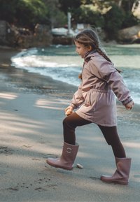 Bottes roses imperméables, leggings marron, et une veste rose clair cintrée à la taille, marchant sur une plage de sable près de l'eau.