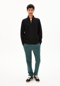 Zwarte ritstrui, teal broek en zwarte sneakers. Model staat met handen in de zakken op een effen witte achtergrond.