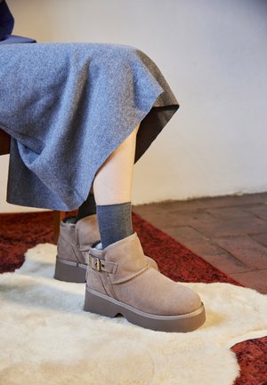 Botas de tobillo de ante color marrón claro con un acento de hebilla, suela de plataforma gris resistente y un interior suave, combinadas con calcetas de canalé grises y una falda gris.