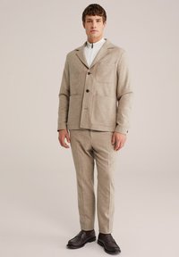 Beige Wollmischjacke mit großen Taschen und Knopfverschlüssen, kombiniert mit passenden schmal zulaufenden Hosen und dunkelbraunen Slippern.