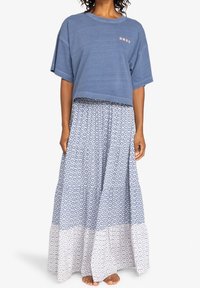 Mujer con una camiseta azul corta y una larga falda azul y blanca con estampado, de pie descalza sobre un fondo blanco.