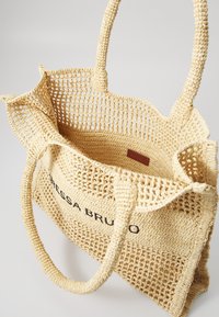 Geweven rieten tote bag in lichtbeige met twee stevige handgrepen, heeft een open netweefsel, merketiket aan de binnenkant en een ruime binnenkant.