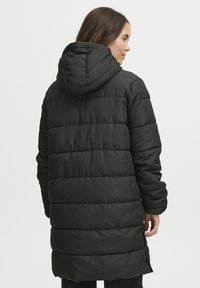 Lungo cappotto piumino nero con cappuccio, caratterizzato da sezioni a cubetto orizzontali e spiacchi laterali per facilitare i movimenti. Texture liscia e finitura opaca.