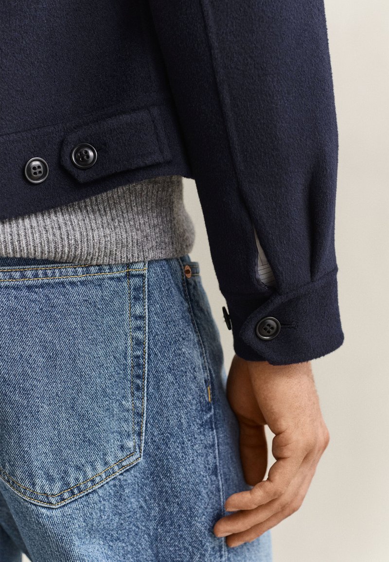 Primo piano di una persona che indossa jeans blu, un maglione grigio a maglia e una giacca blu scuro con polsini abbottonati e una linguetta laterale.
