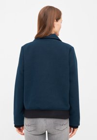 Giacca in pile blu navy con colletto, maniche svasate e orlo nero a coste. Texture liscia e design semplice, vista da dietro.