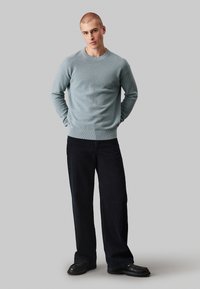 Lichtblauwe gebreide sweater met een ronde hals, gecombineerd met donkere wijde broekspijpen en zwarte schoenen, staand tegen een neutrale achtergrond.