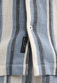 Tissu en maille gaufrée avec des rayures verticales bleu, noir et crème, avec un bouton et une étiquette de la marque "Petrol Industries".