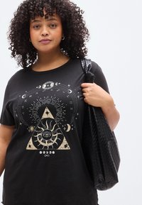 Černé bavlněné tričko s potiskem zlatých geometrických a nebeských motivů, včetně slunce uvnitř trojúhelníku a různých symbolů. Nošeno s černou taškou.