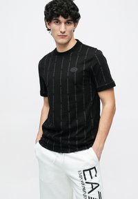 Fiatal férfi göndör hajjal, fekete EA7 Emporio Armani pólót és fehér nadrágot visel nagy EA7 logóval, kezei a zsebében.