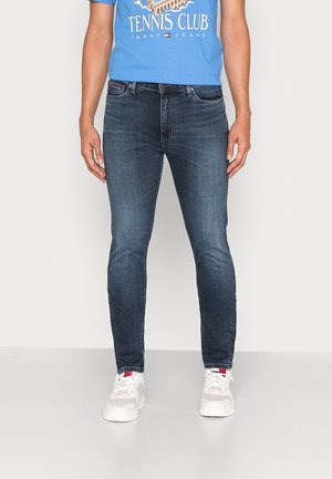 Mand iført mørkeblå slim-fit jeans, hvide sneakers og en blå T-shirt med teksten "Tennis Club", står foran en ensfarvet baggrund.
