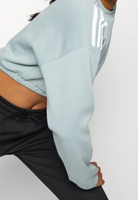 Felpa azzurra chiara con vestibilità rilassata e maniche lunghe. Presenta strisce bianche sulle spalle e una texture morbida. Abbinata a pantaloni neri con cordino.