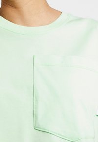 Světle zelené bavlněné tričko s kulatým výstřihem, krátkými rukávy a levým náprsním kapsou s kontrastním šitím.