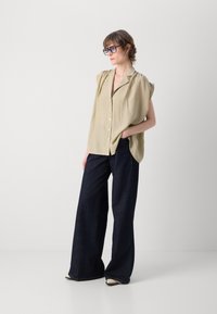 Beige oversized skjorte med skulderfolder, knappelukking, paret med mørke vide jeans og kremfargede sko; solbriller fullfører stilen.