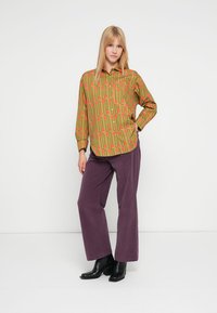 Green button-up shirt with a red and yellow abstract pattern, paired with purple wide-leg pants and black ankle boots.