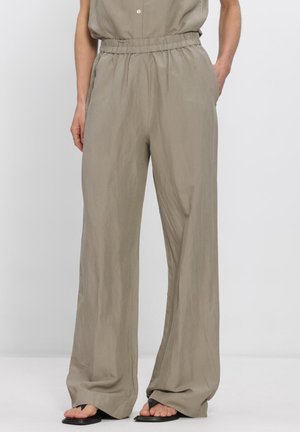 Beige Hosen mit weitem Bein und elastischem Bund, getragen mit schwarzen Riemchensandalen, von der Taille bis zu den Füßen an einer Person vor einfachem Hintergrund gezeigt.