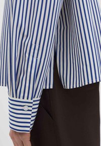 Weißes Hemd mit blauen vertikalen Streifen, ausgestattet mit einem geknöpften Manschetten, seitlichen Schlitzen und einem dezenten Taschen-Detail. Der Stoff wirkt glatt und frisch.