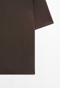 Gros plan sur le tissu d'un T-shirt marron foncé à manches courtes, montrant l'ourlet et les bords des manches sur fond blanc.