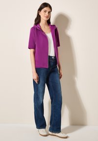 Lila zip-up hoodie med korta ärmar, kombinerad med blå vida jeans och vita sneakers, med en enkel design och synliga sömmar.