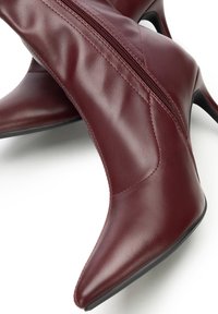 Pár bordó kožených kotníkových bot se špičatou špičkou, bočním zipem a tenkými vysokými podpatky na bílém pozadí.