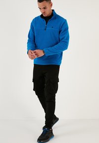 Pullover in pile blu royal con zip a quarter, dettagli neri e un piccolo logo sul petto; indossato con pantaloni cargo neri e sneaker nere.