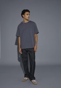 T-shirt grigio oversize, maniche corte, abbinato a jeans in denim scuro. Le scarpe presentano sneakers beige con lacci e righe bianche.