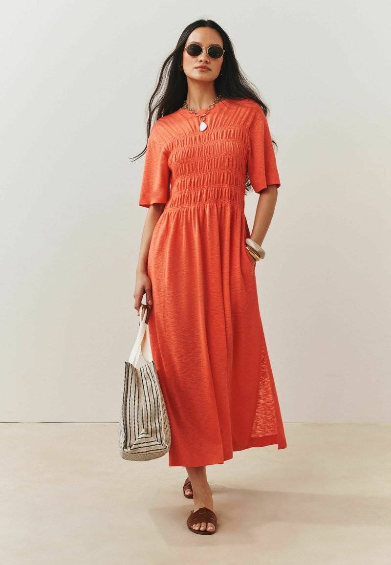 Femme portant une robe midi orange et des lunettes de soleil, portant un sac fourre-tout rayé, et marchant à l'intérieur sur un sol et un mur clairs.