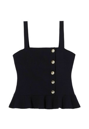 Femme aux longs cheveux foncés portant un haut bleu marine à boutons avec un ourlet à volants, un pantalon large crème, des sandales noires et un foulard bleu à motifs.