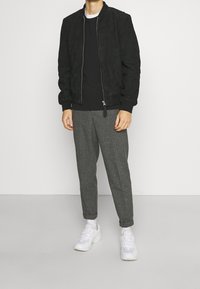 Schwarze Wildleder-Bomberjacke mit Reißverschluss, schwarzer Pullover, grau karierte Hose und weiße Sneakers vor einem neutralen Hintergrund.