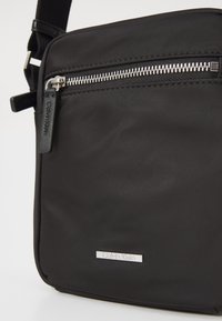 Sac bandoulière noir en nylon avec un design plat, fermeture éclair argentée et plaque logo Calvin Klein. Bandoulière réglable pour un transport facile.
