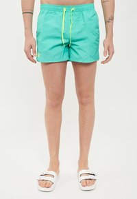 Shorts de bain turquoise avec taille élastique, cordon jaune, et poches latérales. Stylé avec des sandales blanches à glisser. Texture lisse.
