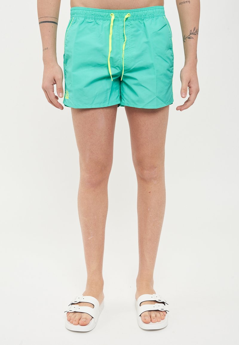 Shorts de bain turquoise avec taille élastique, cordon jaune, et poches latérales. Stylé avec des sandales blanches à glisser. Texture lisse.
