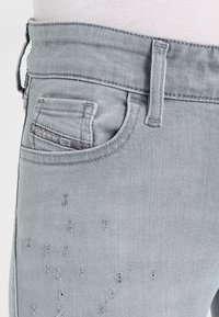 Jeans de denim gris claro con cinturilla, un bolsillo trasero con detalles de costura y acentos desgastados en la parte delantera.