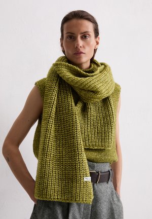 Donna che indossa un top senza maniche in maglia verde oliva e sciarpa abbinata, abbinati a pantaloni grigi e cintura nera, in piedi contro un muro bianco.