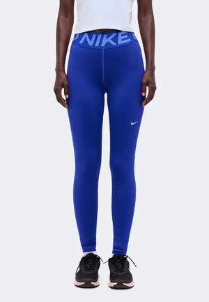 Persoon die felblauwe Nike legging draagt met wit swoosh-logo op de dij en zwarte sportschoenen met witte zolen.