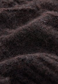 Mørkebrun tekstureret stof med en blød, fnugget overflade, der har ujævne, bølgede folder og en blanding af mørke og lyse fibre.