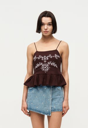 Femme aux cheveux courts et foncés portant un top spaghetti marron foncé avec une broderie florale blanche et une jupe en jean asymétrique cloutée.